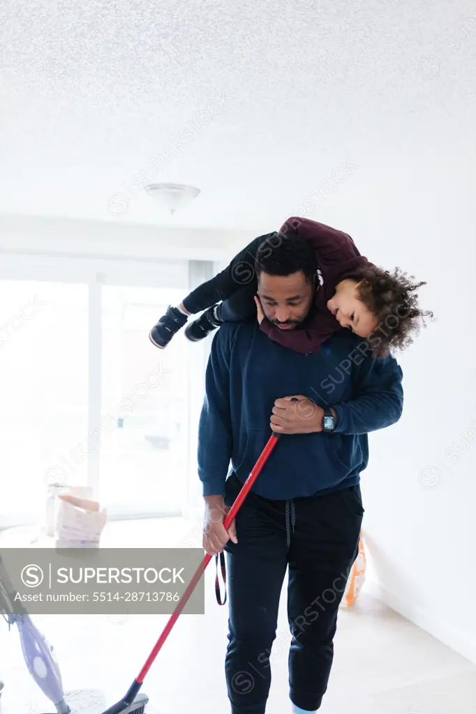 Black father cleaning the house, sweeping, and toddler on shoulders