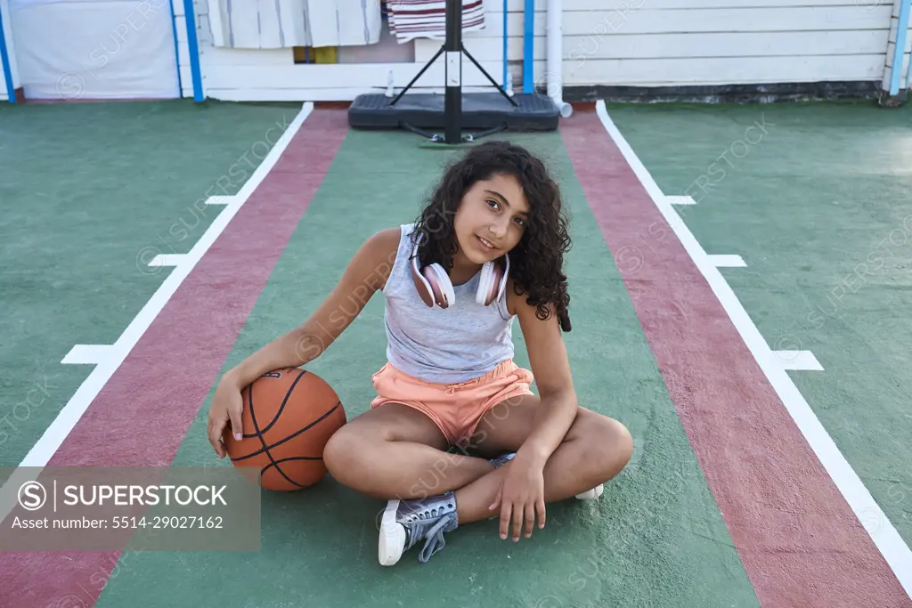 A girl sitting with a basketball facing the camera