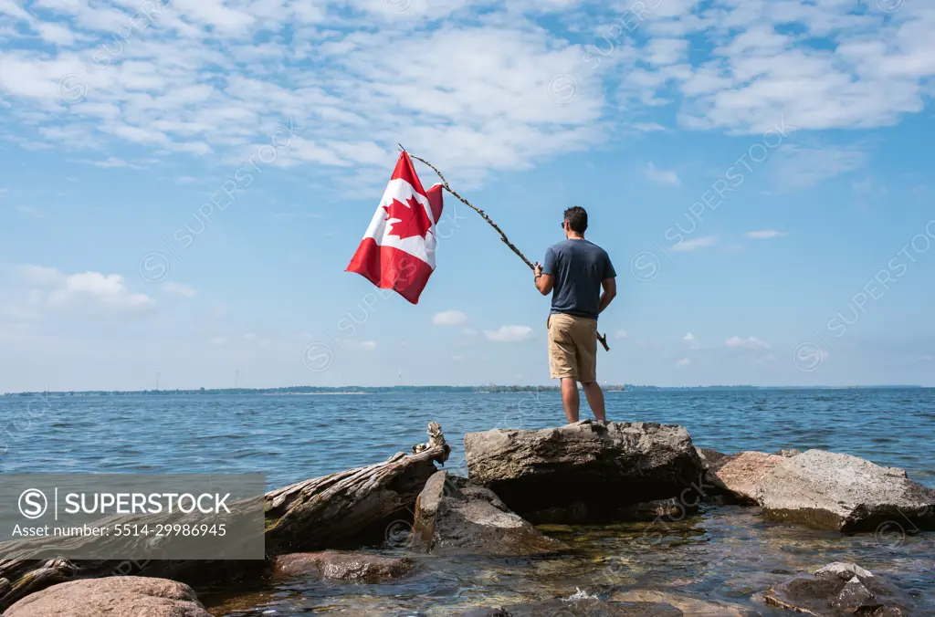 Man holding Canada flag on rocky shore of a lake on a summer day.