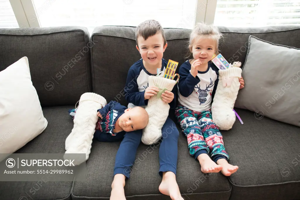 Three kids sit on couch holding their Christmas stockings