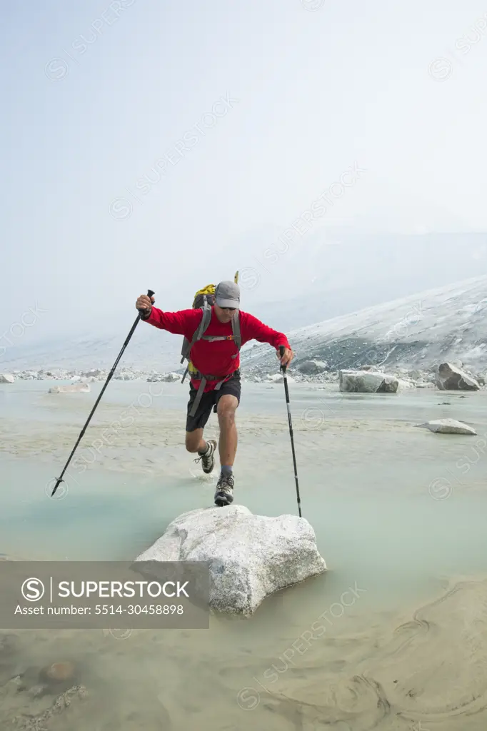 Backpacker leaps over stream onto boulder near glacier.