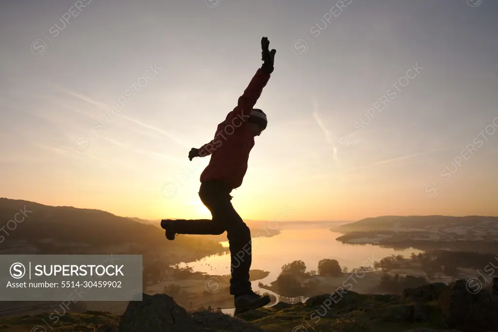 A man jumping for joy at sunrise over Lake Windermere from Todd Crag above Ambleside, Lake District, UK.