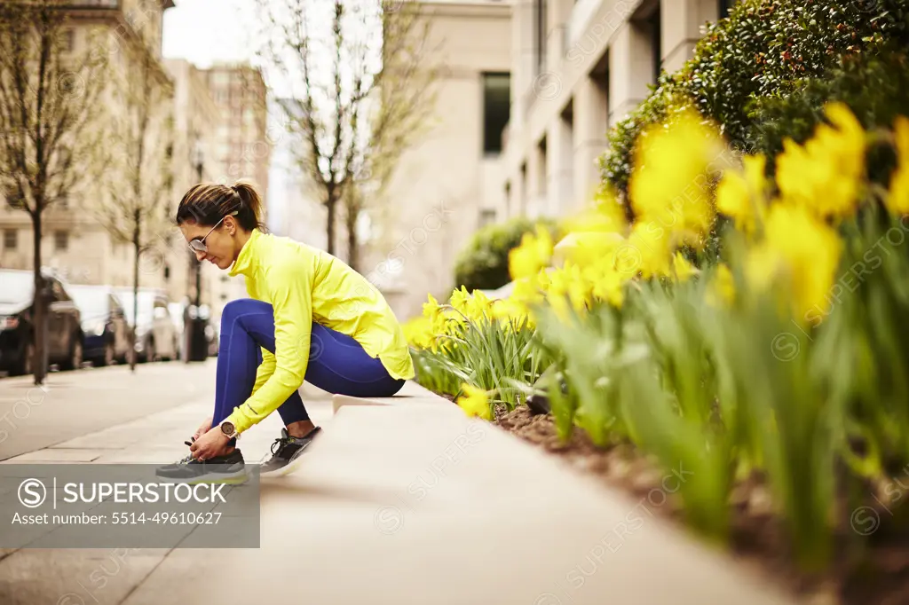 An woman ties her sneakers near some daffodils on a city street.