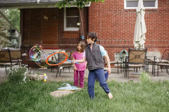 Two children create giant bubbles in yard with a bubble wand