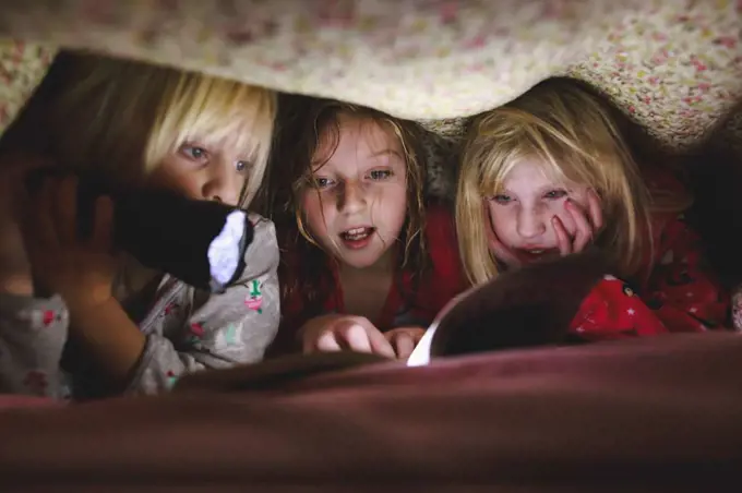 Three little girls reading book under blanket with flashlight