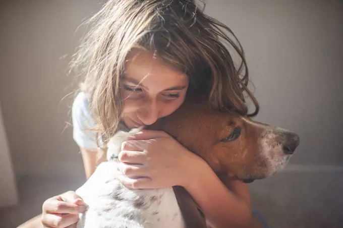 Happy Tween Girl hugging her Basset hound dog inside near a window