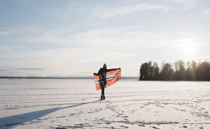 woman walking across a frozen lake holding a Norwegian flag at sunset