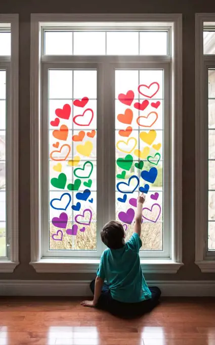 Boy sitting on floor by a window covered in a rainbow of paper hearts.