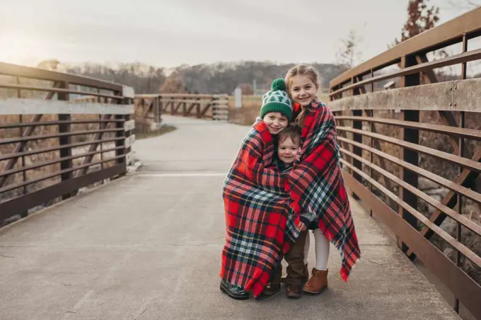 Siblings wrapped in Christmas plaid blanket on bridge at sunset