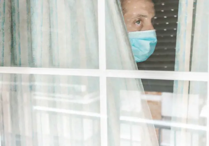 Photo of a Middle-aged white man with medical face mask looking out of the window during coronavirus quarantine