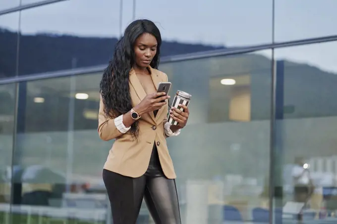 Portrait of black woman wearing a brown suit talking on the phone and holding a container with coffee in the street