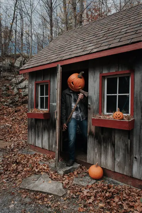 Man wearing scary carved pumpkin head in shed with axe for Halloween.