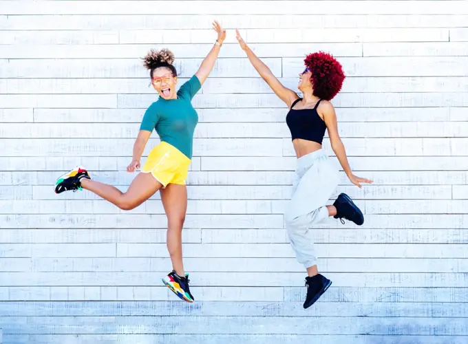 Two latin women with afro hair jumping and high five on white wall.