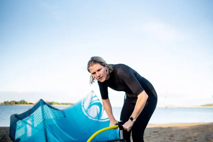 A woman in wetsuit pumps up a kiteboarding kite on a beach