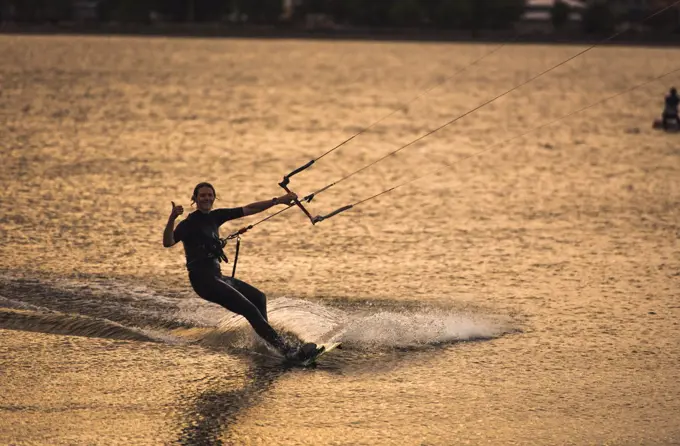 A woman giving up a thumbs up while kiteboarding during late day