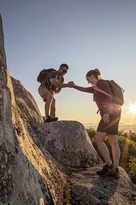 African American male helps female hiker up ledge on mountain in Maine