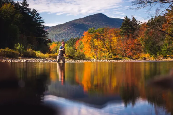 Fly-fisherman casting in river with foliage and reflections