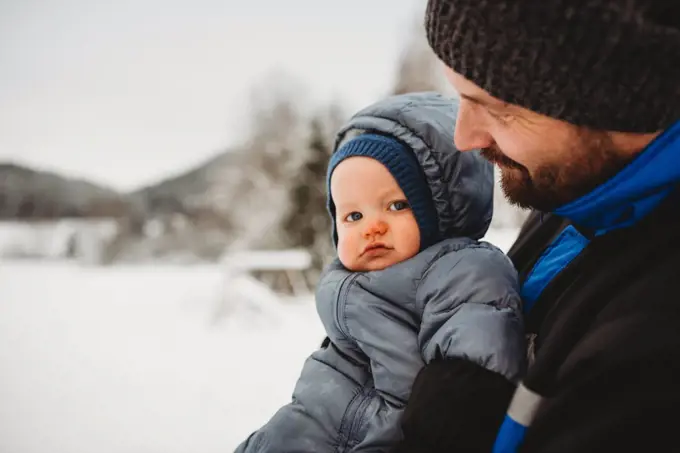 close up of Dad holding baby wearing snowsuit in winter with snow