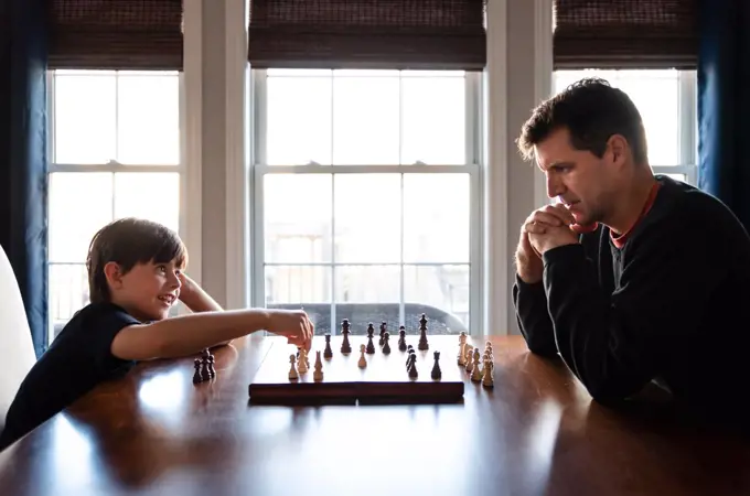 Father and son sitting at a table indoors playing a game of chess.