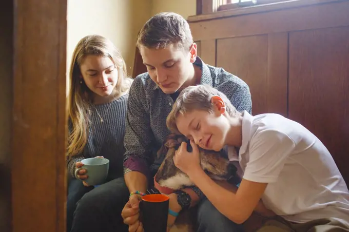 Three happy siblings cuddle their dog in beautiful golden window light