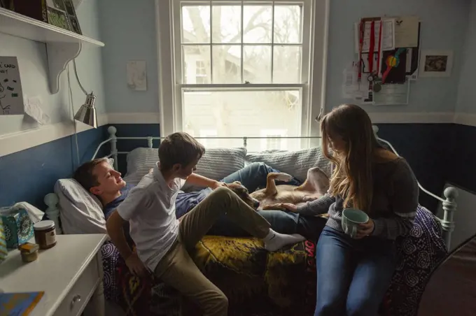 three siblings sit on a bed cuddling with a cute family dog in bedroom