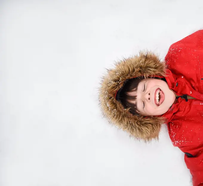 Overhead of happy boy in red coat with furry hood laying in the snow.