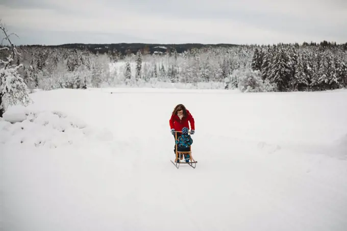 Mother pushing child on sleigh in snow in woods on cold winter day