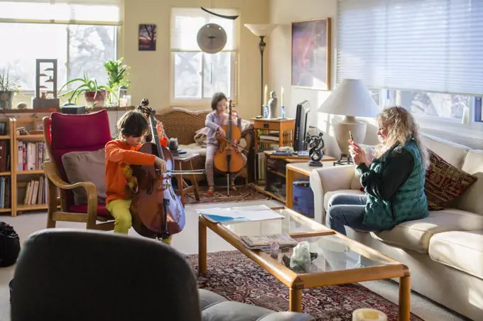 Two children perform cello at home and grandmother records with phone