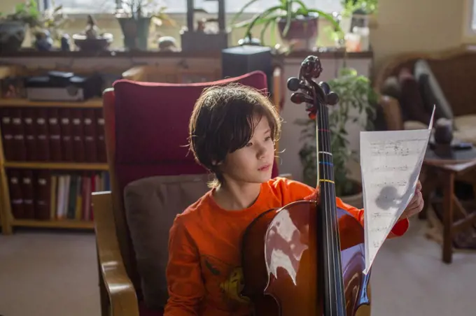 A boy sits in beautiful light with a cello reading sheet music