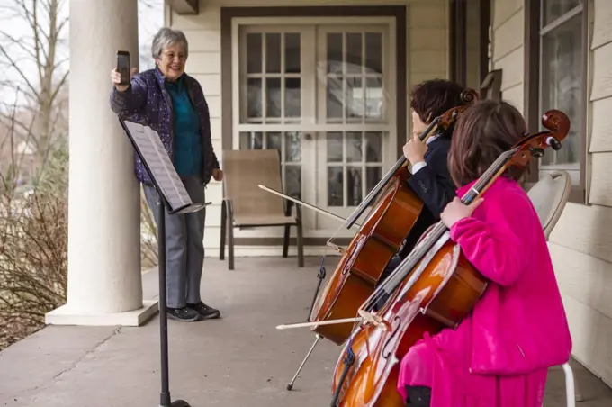 An elderly woman records two children giving cello concert on porch