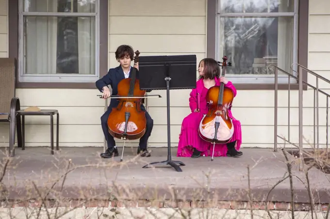 A tween boy sits on porch playing cello in suit while sister watches