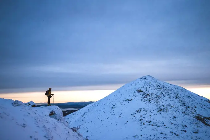 Man stands on cliff in snow in winter looking at distant mountain