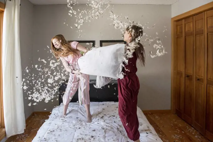 Two happy young girls having a feather pillow fight on the bed.