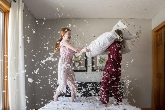 Two happy young girls having a feather pillow fight on the bed.