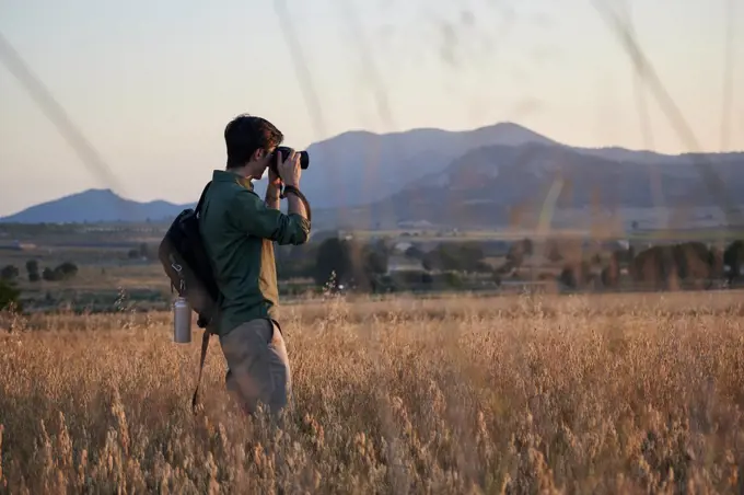 Man photographer with a backpack takes a photo in a meadow at sunset