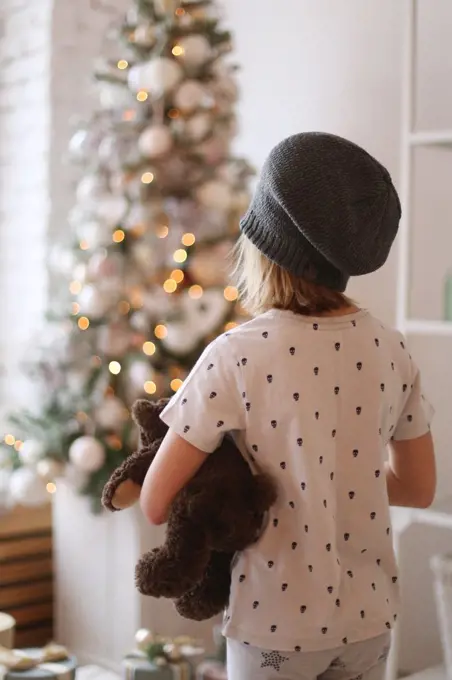 a child in a hat stands in front of a christmas tree