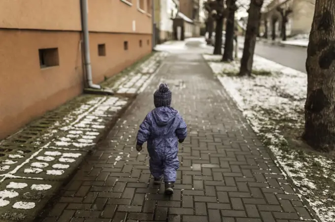 Little boy in blue onesie and winter hat walking on pavement