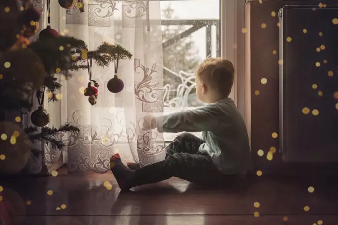 Toddler boy sitting on the floor by the window next to a christm