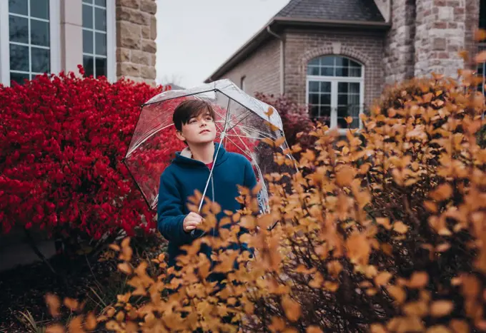 Boy holding umbrella in front of house on a rainy fall day.