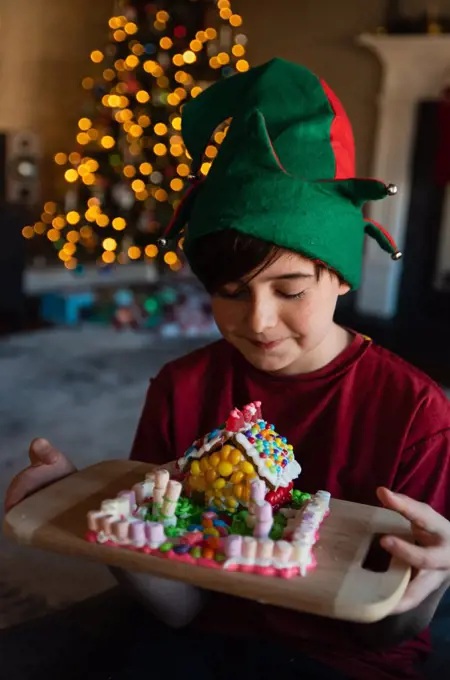 Happy boy in elf hat looking at gingerbread house at Christmastime.