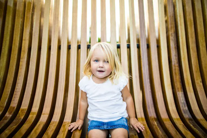 Cute blonde girl sitting on wooden bench at park.