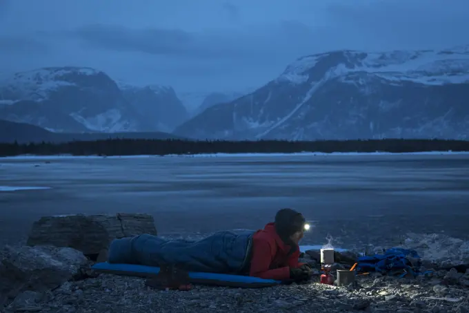 Male hiker preparing food while lying in sleeping bag on mountain at dusk