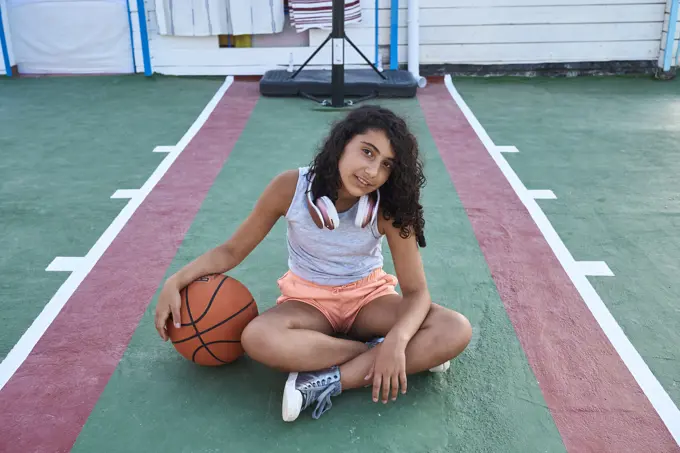 A girl sitting with a basketball facing the camera