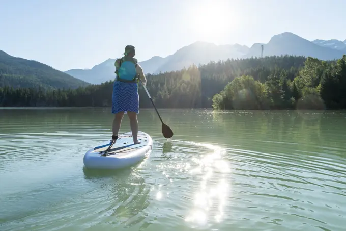 Back view of barefoot woman in dress and life vest riding SUP board on calm lake water on sunny summer day in mountains in British Columbia, Canada