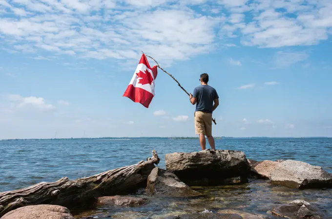 Man holding Canada flag on rocky shore of a lake on a summer day.