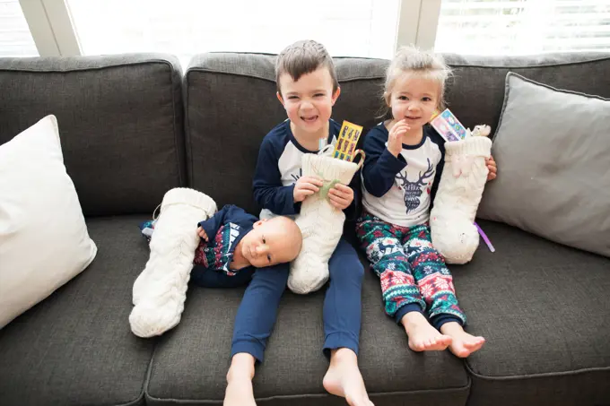 Three kids sit on couch holding their Christmas stockings