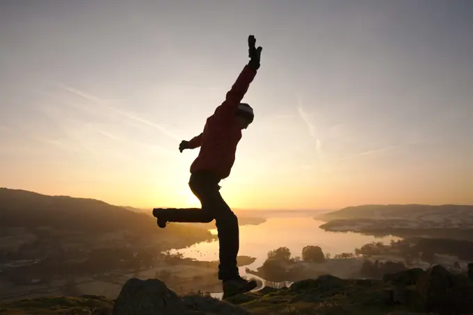 A man jumping for joy at sunrise over Lake Windermere from Todd Crag above Ambleside, Lake District, UK.