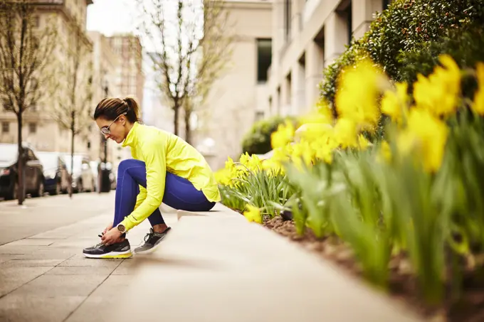 An woman ties her sneakers near some daffodils on a city street.