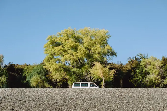 Isolated white van on rocky beach with calm green trees in background