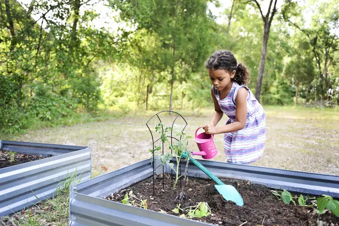 Girl watering plants in a garden bed with a pink watering can ou